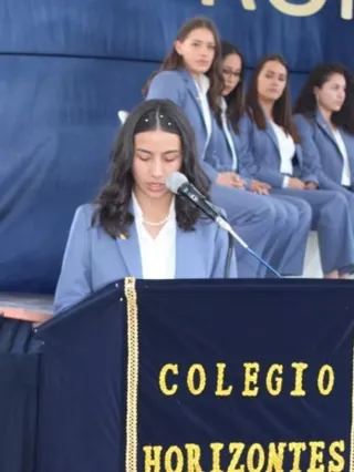 Una alumna del Colegio Horizontes lee desde el atril en un acto institucional, con sus compañeras de fondo.