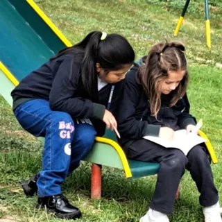 Dos alumnas del Colegio Horizontes leen juntas sentadas en un tobogán verde del área de juegos sobre el césped.