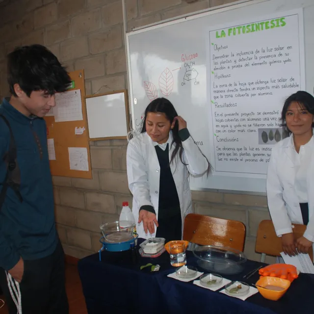 Dos alumnas del Colegio Horizontes, con bata blanca, muestran su experimento sobre la fotosíntesis a un estudiante durante la feria multidisciplinaria de secundaria.