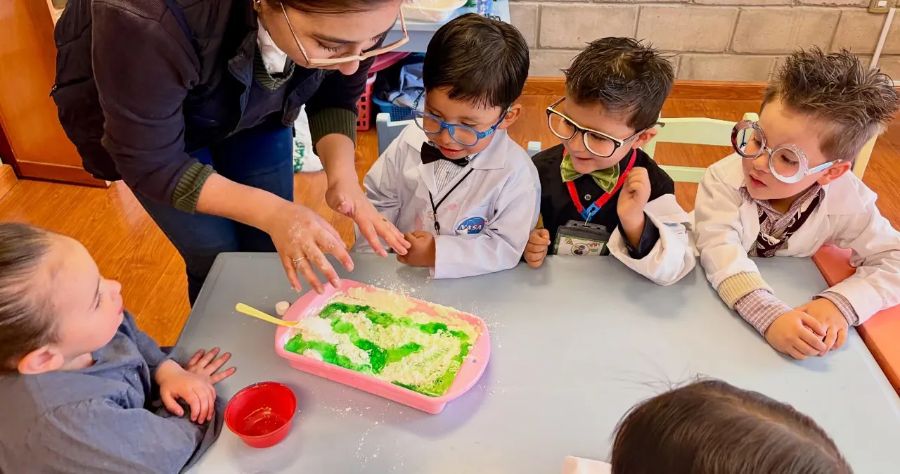 Una profesora del Colegio Horizontes guía a un grupo de estudiantes de nivel inicial, vestidos de científicos, mientras realizan un experimento con espuma verde sobre la mesa.