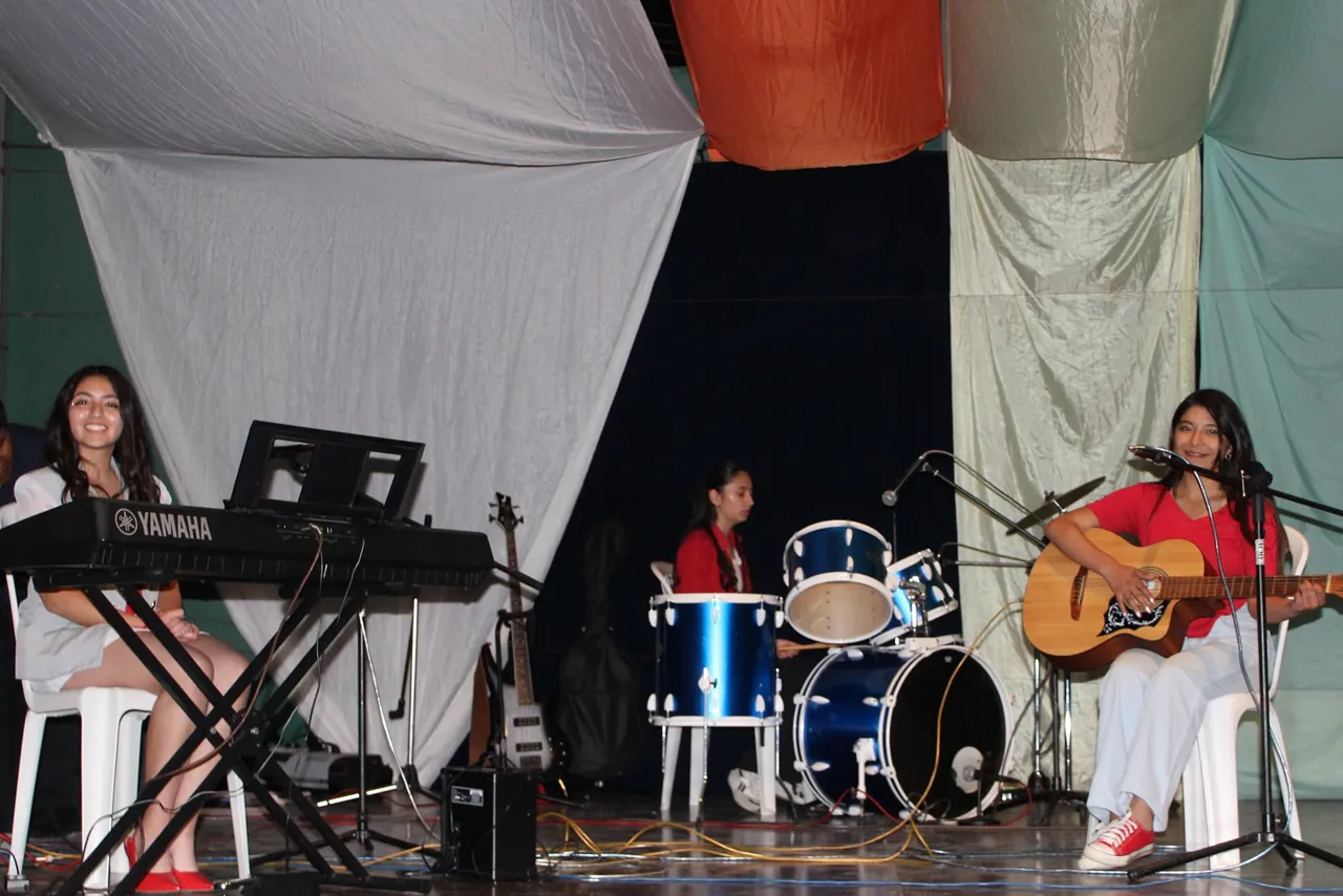 Tres alumnas del Colegio Horizontes tocan el teclado, la batería y la guitarra en un ensamble musical sobre el escenario.