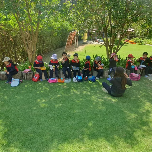Niños y niñas del Nivel Inicial meriendan sentados en troncos bajo los árboles del jardín del Colegio Horizontes.