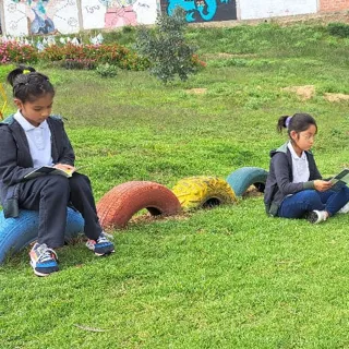 Dos alumnas del Colegio Horizontes leen un libro sentadas sobre los neumáticos coloridos del área de juegos, rodeadas de césped.