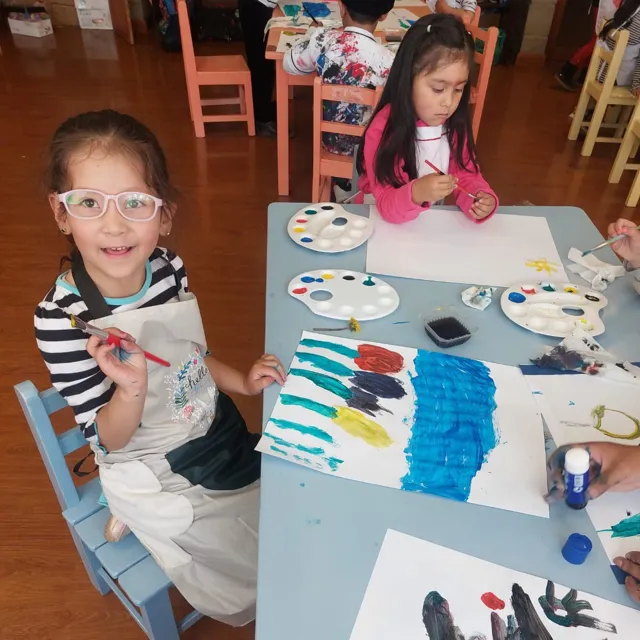 Una alumna del Colegio Horizontes sonríe con un pincel en la mano junto a una pintura de colores, mientras otra compañera pinta al fondo en el aula de artes plasticas.