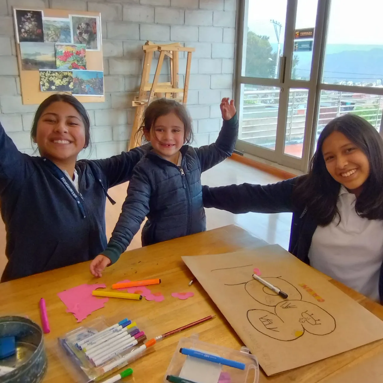 Dos alumnas mayores y una niña pequeña del Colegio Horizontes levantan los brazos sonriendo en el aula de artes plásticas, con un caballete y láminas de pintura de fondo.
