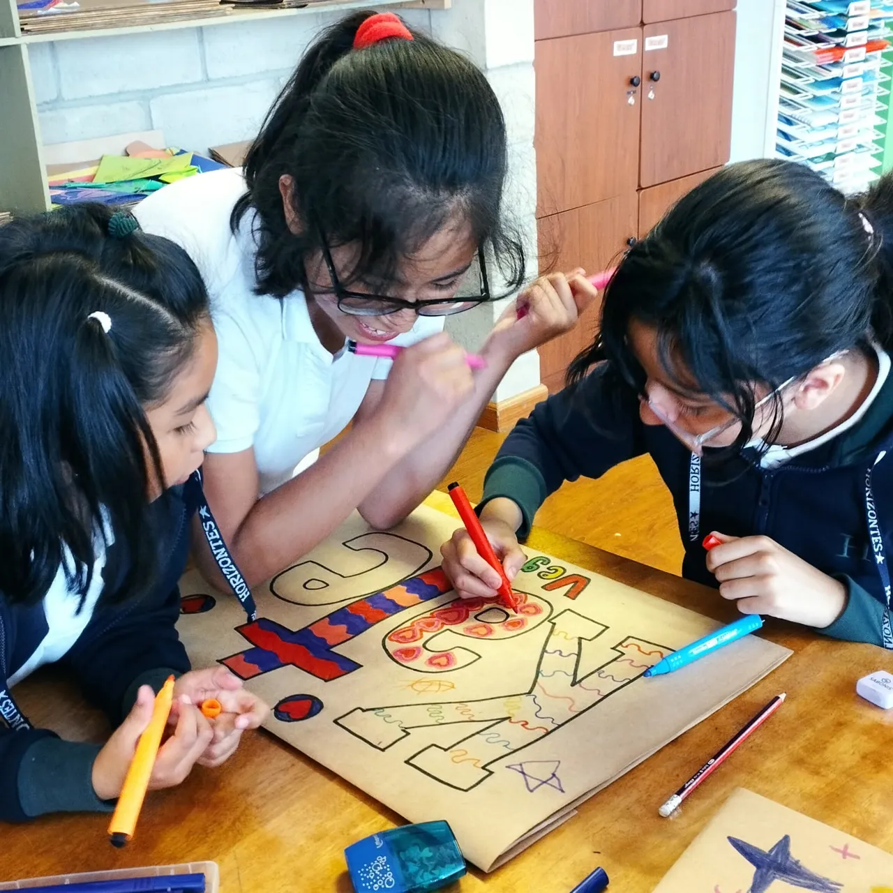 Tres alumnas del Colegio Horizontes colorean juntas un cartel grande con marcadores en la clase de arte.