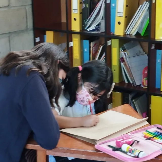 Una alumna del Colegio Horizontes escribe en su cuaderno junto a su profesora, frente a una estantería con libros y carpetas de estudio.