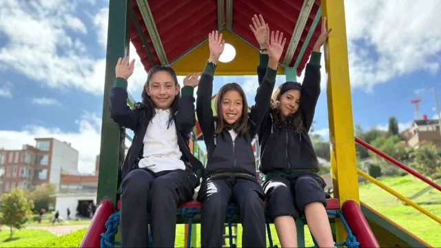 Tres alumnas levantan los brazos sentadas en el tobogán del patio de juegos del colegio
